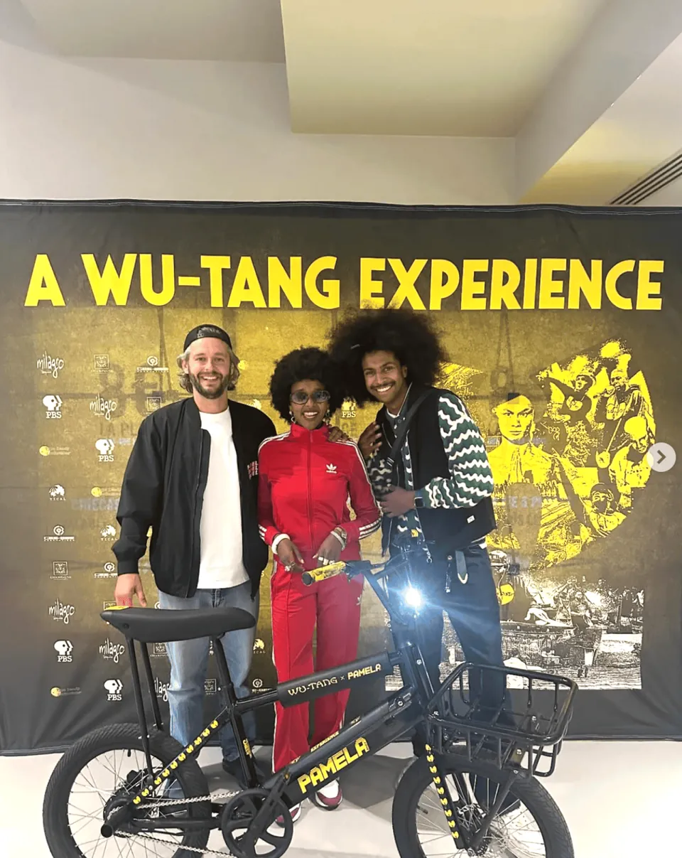 Trois personnes posent derrière un Pamela noir aux détails jaunes, devant l’affiche « A Wu-Tang Experience »